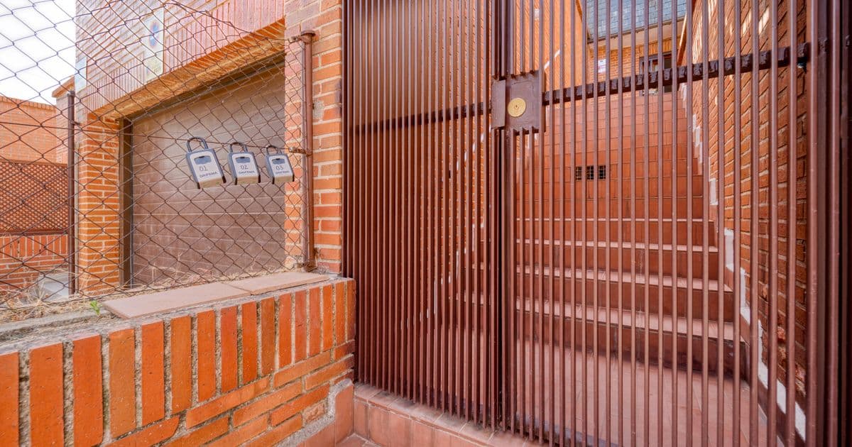 Portões metálicos marrons de barras verticais na entrada de uma casa de tijolo aparente, com degraus de piso cerâmico ao fundo