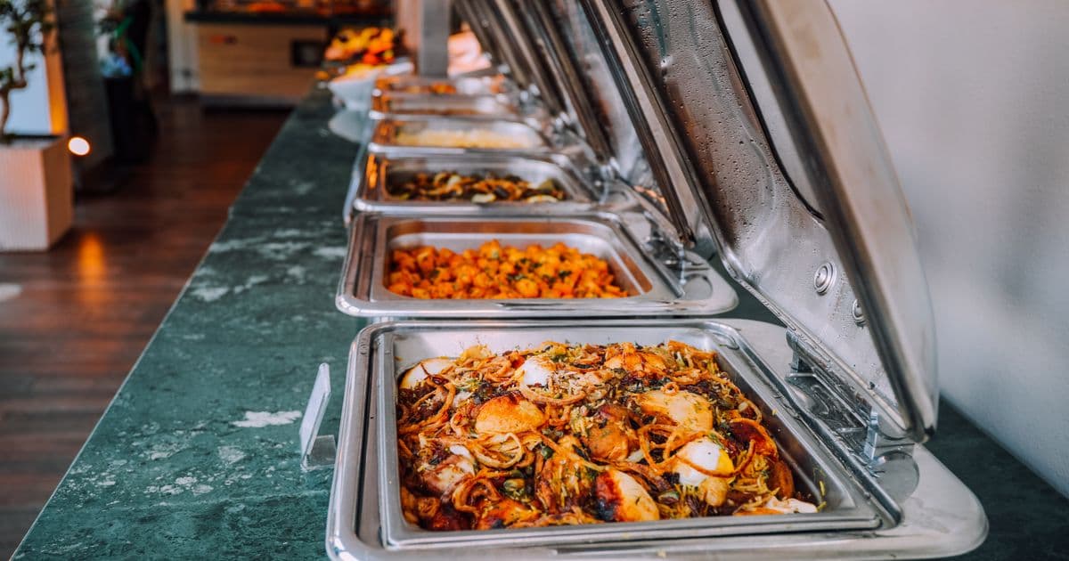 Fila de rechauds de inox sobre balcão de mármore verde, com pratos quentes servidos em buffet de evento