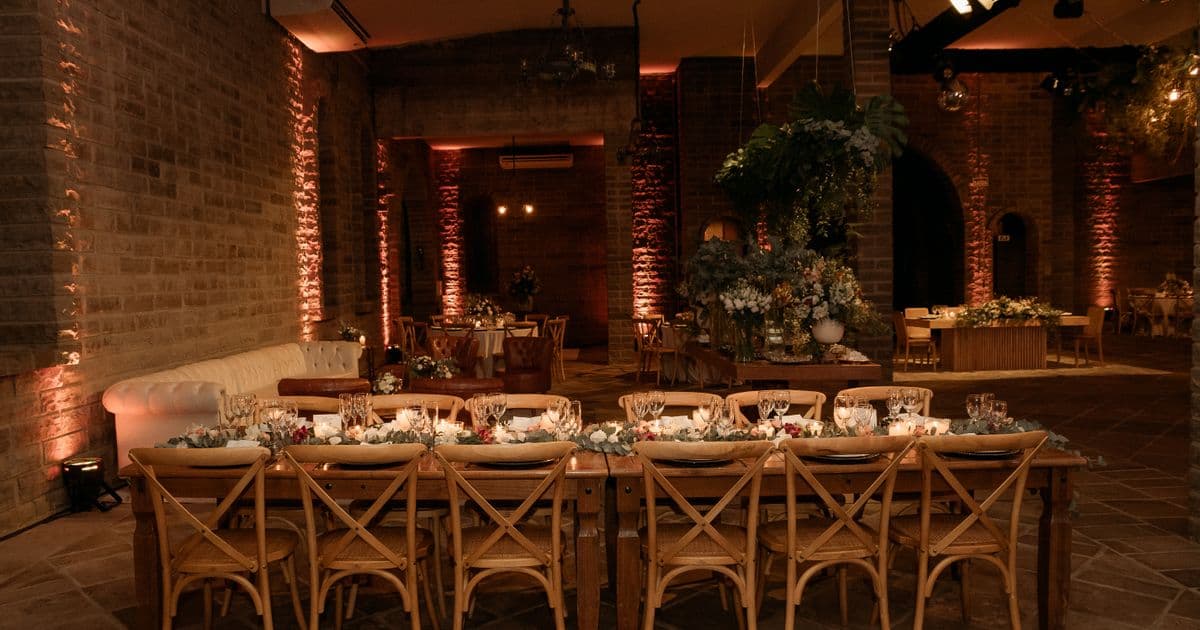 Salão de casamento montado antes da festa, com mesa comprida de madeira, velas acesas, caminho de folhagem e cadeiras cross-back