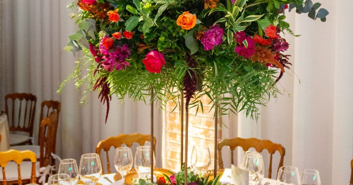 Arranjo floral alto em tons de laranja e rosa sobre mesa posta de casamento, com taças e cadeiras de madeira